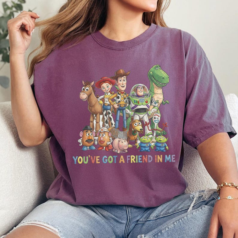 May include: A maroon t-shirt featuring a colorful graphic of characters from the animated film Toy Story. The phrase "YOU'VE GOT A FRIEND IN ME" is printed in rainbow colors below the characters. The shirt has short sleeves.