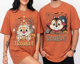 May include: Two rust-colored t-shirts featuring cartoon chipmunks in Halloween costumes. The left shirt shows a mummy chipmunk with the text TROUBLE. The right shirt shows a vampire chipmunk with the text DOUBLE, surrounded by bats, webs, and pumpkins.