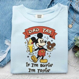 Puede incluir: Camiseta azul claro con un gr&aacute;fico de Mickey Mouse sosteniendo un batido y una rosquilla. El texto "DAD TAX" est&aacute; encima de Mickey, y "IF I'M BUYIN' I'M TRYIN'" debajo. La camiseta est&aacute; doblada sobre una superficie de mezclilla.