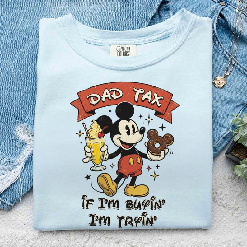 Puede incluir: Camiseta azul claro con un gr&aacute;fico de Mickey Mouse sosteniendo un batido y una rosquilla. El texto "DAD TAX" est&aacute; encima de Mickey, y "IF I'M BUYIN' I'M TRYIN'" debajo. La camiseta est&aacute; doblada sobre una superficie de mezclilla.