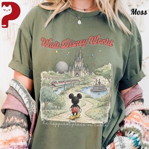 May include: Olive green t-shirt featuring a colorful illustration of Walt Disney World. The graphic includes the castle, Spaceship Earth, a monorail, and Mickey Mouse. Text reads "Walt Disney World" and "The happiest place on earth."
