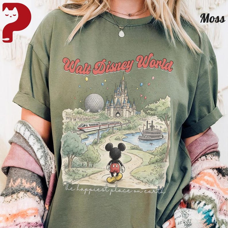 May include: Olive green t-shirt featuring a colorful illustration of Walt Disney World. The graphic includes the castle, Spaceship Earth, a monorail, and Mickey Mouse. Text reads "Walt Disney World" and "The happiest place on earth."