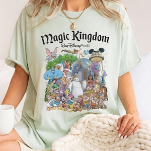 May include: Light green t-shirt featuring a colorful graphic of Disney characters, including Mickey Mouse, Aladdin, and Baymax. The text "Magic Kingdom Walt Disney World" is printed above the characters. The shirt has short sleeves and a crew neck.