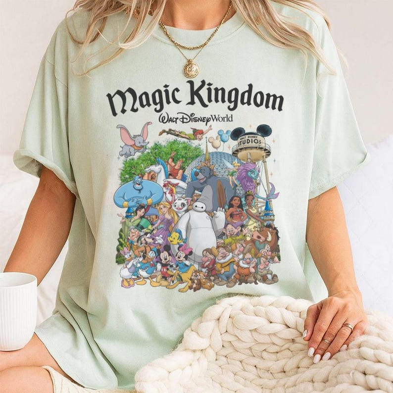 May include: Light green t-shirt featuring a colorful graphic of Disney characters, including Mickey Mouse, Aladdin, and Baymax. The text "Magic Kingdom Walt Disney World" is printed above the characters. The shirt has short sleeves and a crew neck.