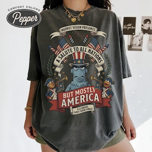 May include: Dark gray Comfort Colors t-shirt featuring a Muppet Vision graphic. The design includes the text "Muppet Vision Presents, A Salute to All Nations, But Mostly America, A Glorious Three Hour Finale". The shirt has a distressed vintage look.