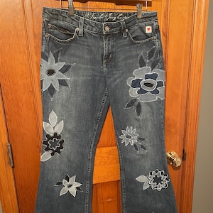 May include: Blue denim jeans with a wide leg and floral embroidery in shades of blue and black. The jeans have a small red heart patch on the back pocket.
