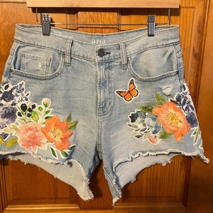 May include: Light blue denim shorts with floral and butterfly appliques. The shorts have a frayed hem and are labeled "ana".