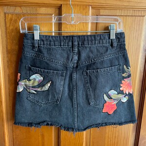 May include: A black denim mini skirt with a frayed hem and floral embroidery. The skirt has a back pocket and a zip closure.