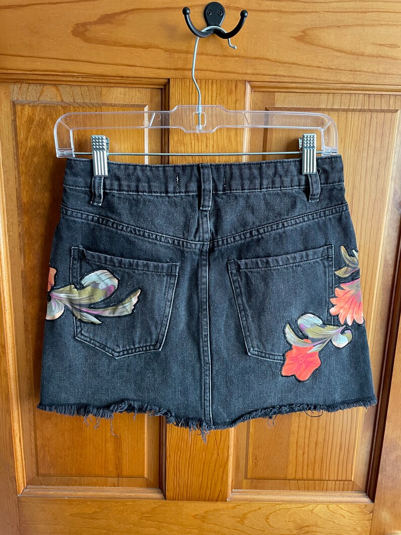 May include: A black denim mini skirt with a frayed hem and floral embroidery. The skirt has a back pocket and a zip closure.