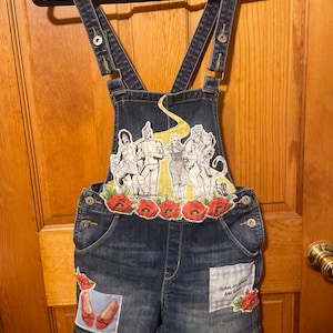May include: Blue denim overalls with a yellow brick road and characters from the Wizard of Oz printed on the front. The overalls have red poppy flowers and the text "There's no place like home" on the legs.