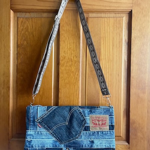 May include: A denim crossbody bag with a flap closure, featuring a patch pocket and a Levi Strauss & Co. label. The bag has a patterned strap and is hanging on a wooden door. The bag is made from recycled denim.