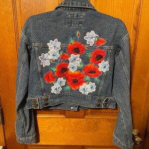 May include: A denim jacket with a floral design on the back. The jacket features a cropped fit, button closures, and a classic collar. The floral design includes red poppies and blue flowers.
