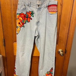 May include: Light wash denim jeans with floral embroidery in red, orange, and yellow. The jeans feature embroidered flowers on the front and bottom hems, with a red and yellow trim and yellow pom-poms. The back pocket has a red patterned fabric.