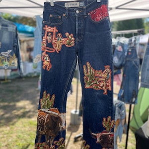 May include: Dark wash denim jeans with colorful embroidered western-themed designs. The jeans feature images of cowboys, cacti, and longhorn cattle. A red bandana detail is visible on the right pocket.