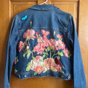 May include: A blue denim jacket with embroidered flowers, birds, and butterflies. The flowers are pink, white, and green. The birds are a cardinal and a bluebird. The butterflies are blue.