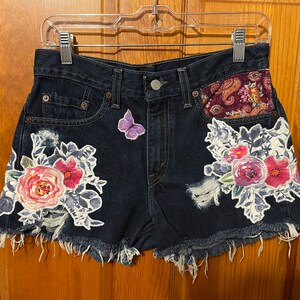 May include: Dark blue denim shorts with floral embroidery and a butterfly applique. The shorts have a frayed hem and a paisley patch on the back pocket.