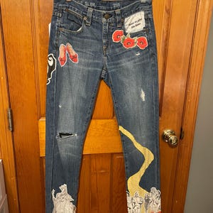 May include: Blue denim jeans with decorative patches featuring red poppies, ruby slippers, and characters from The Wizard of Oz. The jeans have a distressed look with a torn knee and a yellow road design.