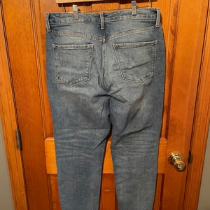 May include: A pair of blue denim jeans with a frayed hem. The jeans have a relaxed fit and are hanging on a wooden hanger.