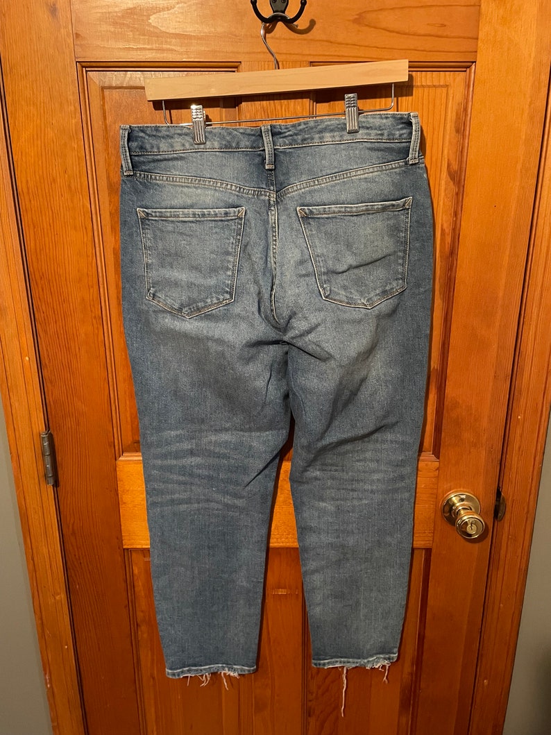 May include: A pair of blue denim jeans with a frayed hem. The jeans have a relaxed fit and are hanging on a wooden hanger.