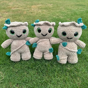 May include: Three crocheted plush toys shaped like a tree with green leaves and brown bodies. The toys have green eyes and a smiling expression.