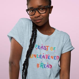 Pode incluir: Uma camiseta azul clara com o texto "FEAST OF UNLEAVENED BREAD" em letras coloridas. A camiseta é usada por uma pessoa com óculos pretos e cabelo comprido trançado.