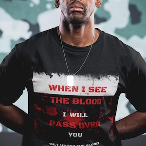 Pode incluir: Camiseta preta com o texto "WHEN I SEE THE BLOOD I WILL PASS OVER YOU" em vermelho e branco. A camisa também apresenta o texto "GET UNDER THE BLOOD EXODUS 12:13". Uma placa de identificação prateada pende de uma corrente.