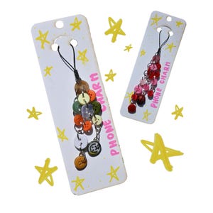 May include: Two phone charms on white cards with yellow star doodles. One charm features a cluster of colorful buttons, the other has red and pink beads. The cards read "PHONE CHARM" in pink.