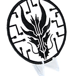 May include: A circular embroidered patch featuring a black dragon-like design on a white background. The patch has a black border and is displayed on a clear plastic stand. The design is intricate and detailed, with sharp lines and curves.