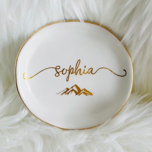 Pu&ograve; includere: Piatto portaoggetti in ceramica bianca con bordo dorato e scritta dorata. Il piatto dice "Sophia" e presenta un design di montagna dorato.
