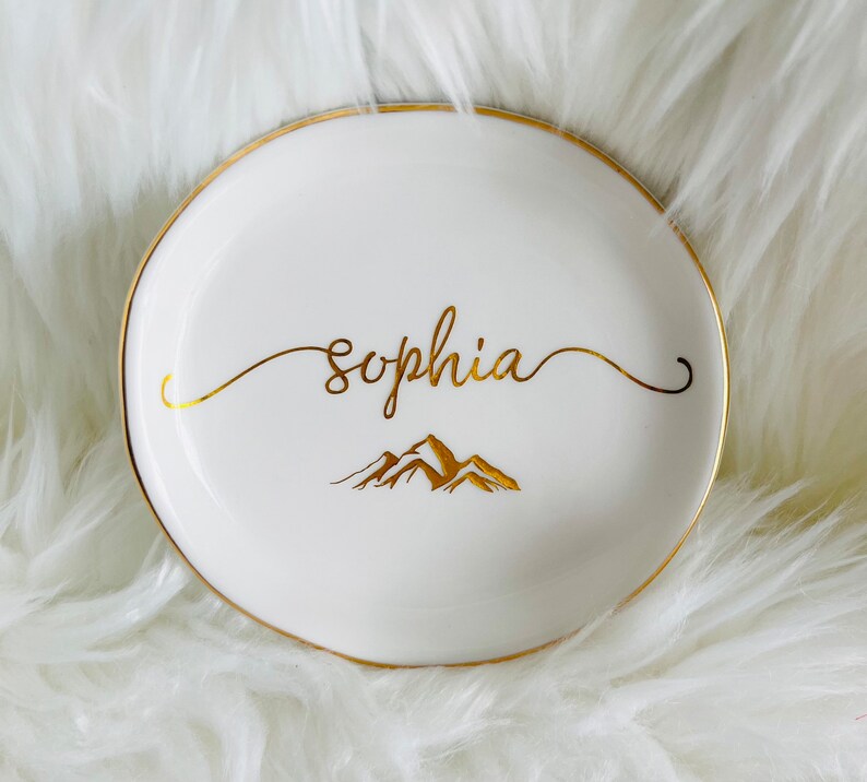 Pu&ograve; includere: Piatto portaoggetti in ceramica bianca con bordo dorato e scritta dorata. Il piatto dice "Sophia" e presenta un design di montagna dorato.