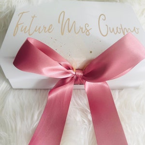 May include: White gift box with a pink satin ribbon tied in a bow. The box has the text "Future Mrs. Cuomo" written in gold lettering.