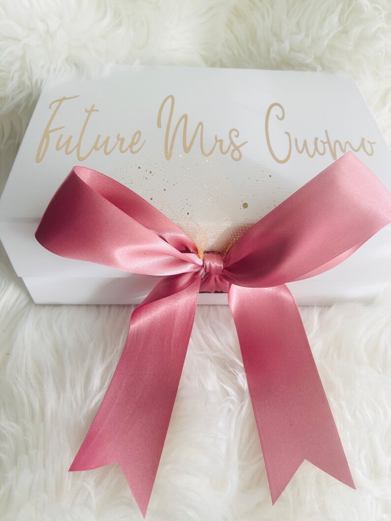 May include: White gift box with a pink satin ribbon tied in a bow. The box has the text "Future Mrs. Cuomo" written in gold lettering.