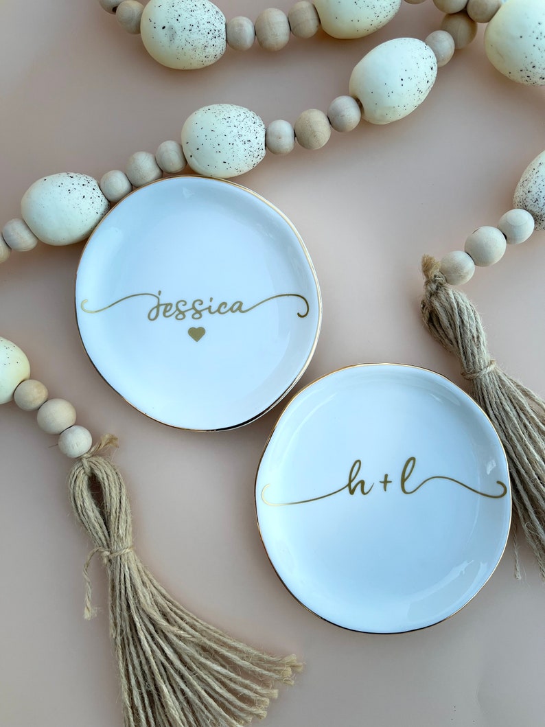 Pu&ograve; includere: Due porta anelli in ceramica bianca con bordo dorato. Un piatto ha il nome "Jessica" scritto in corsivo dorato con un cuore dorato. L'altro piatto ha le iniziali "h+l" scritte in corsivo dorato. Entrambi i piatti sono su uno sfondo rosa chiaro con una ghirlanda di perline di legno con accenti bianchi.