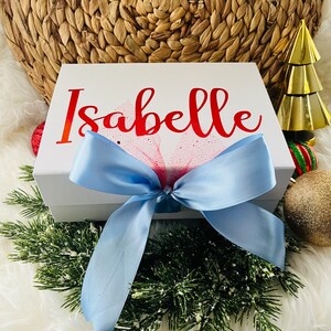 May include: White gift box with a blue ribbon and a red bow. The box is decorated with the name "Isabelle" in red lettering.