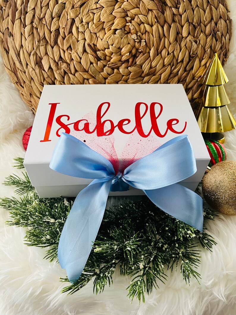 May include: White gift box with a blue ribbon and a red bow. The box is decorated with the name "Isabelle" in red lettering.