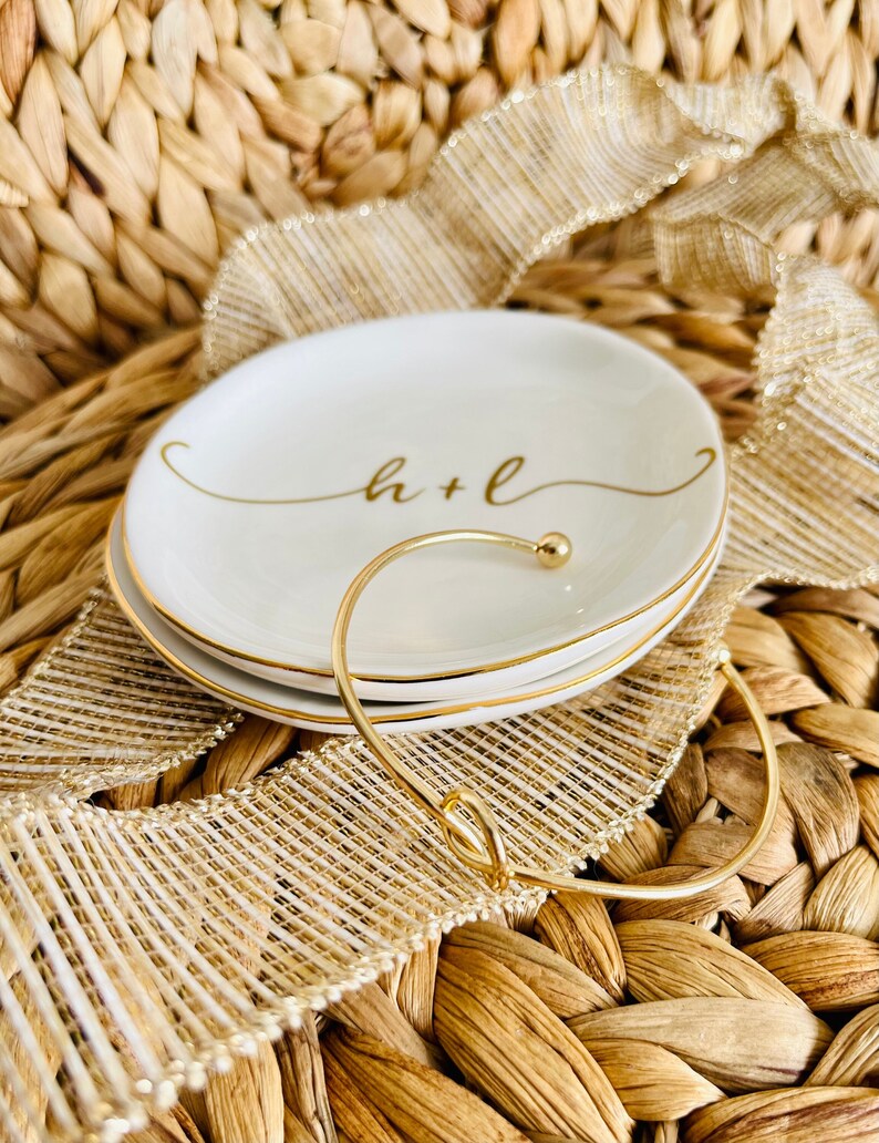 Pu&ograve; includere: Un porta anelli in ceramica bianca con un bordo dorato e l'iscrizione "h+l" in caratteri dorati. Un braccialetto dorato riposa sul piatto.