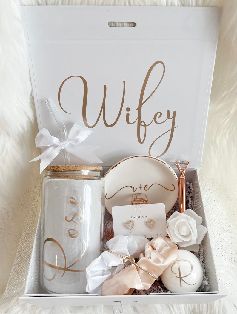 K&ouml;nnte beinhalten: Eine wei&szlig;e Geschenkbox mit einer goldenen Aufschrift, die "Wifey" lautet. Die Box enth&auml;lt eine Vielzahl von Geschenken, darunter ein Glasbecher mit einer goldenen Aufschrift, die "Rose" lautet, eine wei&szlig;e Keramikschale mit einer goldenen Aufschrift, die "cute" lautet, ein Paar goldene herzf&ouml;rmige Ohrringe, eine wei&szlig;e Rose, ein goldenes Haargummi und eine wei&szlig;e Badebombe mit einer goldenen Aufschrift, die "R" lautet.
