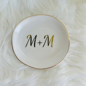 Pu&ograve; includere: Piatto porta anelli in ceramica bianca con bordo dorato e le iniziali "M+M" scritte in lettere dorate.