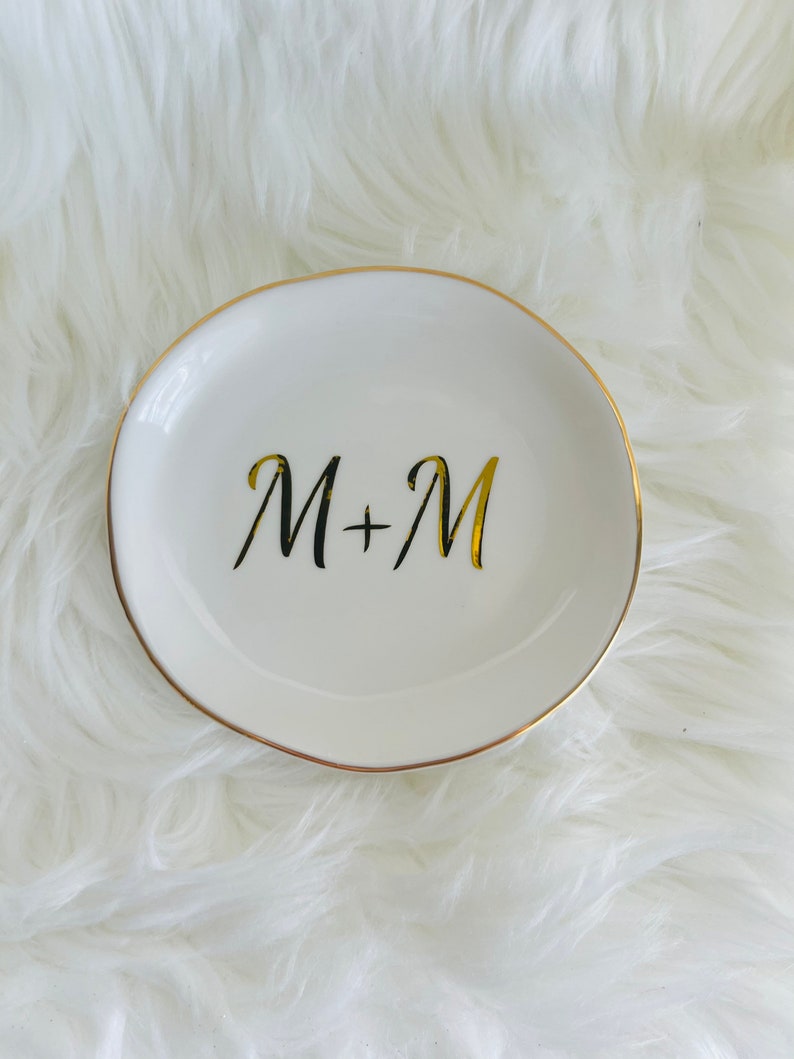 Pu&ograve; includere: Piatto porta anelli in ceramica bianca con bordo dorato e le iniziali "M+M" scritte in lettere dorate.