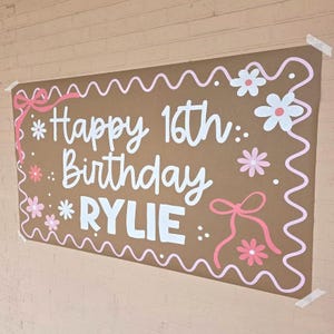 Peut inclure: Une bannière en papier brun avec le texte "Happy 16th Birthday Rylie" en écriture blanche. La bannière est décorée de fleurs roses, de nœuds et d'une bordure rose ondulée. La bannière est fixée à un mur de briques.