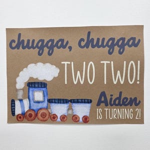 May include: A brown card with a watercolor illustration of a blue train with white smoke. The text "chugga, chugga TWO TWO! Aiden IS TURNING 2!" is written in blue and white.