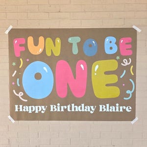 May include: A brown paper banner with the words "FUN TO BE ONE" in colorful bubble letters. Below, it reads "Happy Birthday Blaire" in white. The banner is decorated with confetti and taped to a brick wall.