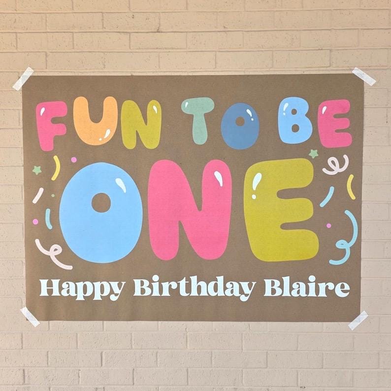 May include: A brown paper banner with the words "FUN TO BE ONE" in colorful bubble letters. Below, it reads "Happy Birthday Blaire" in white. The banner is decorated with confetti and taped to a brick wall.