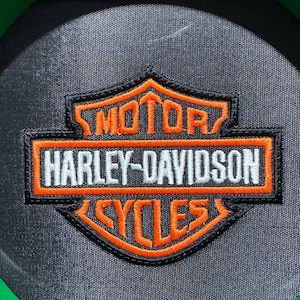 May include: A black and orange Harley-Davidson Cycles patch. The patch features the Harley-Davidson logo with the words "MOTOR HARLEY-DAVIDSON CYCLES" in white and orange lettering. The patch is on a black background and has a green border.