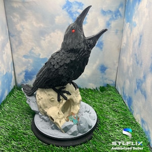 May include: A black raven with red eyes perched on a white skull with a blue and green gemstone. The raven is in a mid-crowing pose with its beak open wide. The skull and raven are on a gray base with a black rim.