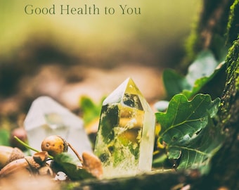 May include: A close-up of a clear, yellow crystal with a pointed top, surrounded by acorns, leaves, and another crystal. The text "Good Health to You" is at the top. The crystals are on a bed of leaves and moss.