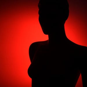 Op de afbeelding: Zwart silhouet van een persoon tegen een levendige rode achtergrond. De vorm bevindt zich in de schaduw, met de omtrek van het hoofd, de schouders en de torso duidelijk zichtbaar. De belichting creëert een sterk contrast tussen de donkere figuur en de heldere rode achtergrond.