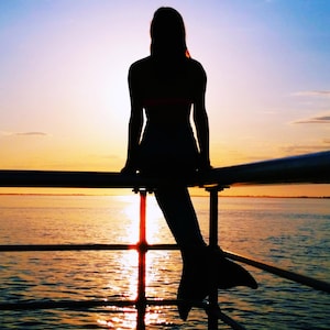 Puede incluir: Silueta de una persona con cola de sirena sentada en una barandilla, mirando la puesta de sol. El cielo transiciona de azul a naranja y amarillo, reflejándose en el agua. La cola de sirena es oscura y la barandilla es de metal oscuro.