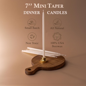 May include: A display of white 7-inch mini taper dinner candles. The candles are on a wooden holder with a gold base. The image includes text that says "Small Batch", "All Natural", "Non Toxic", and "100% USA Beeswax".