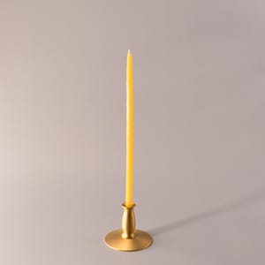 May include: A tall, slender yellow taper candle standing upright in a matte gold metal candlestick holder. The candle has a smooth, cylindrical shape with a pointed tip, and the holder features a wide, circular base and a tapered neck.